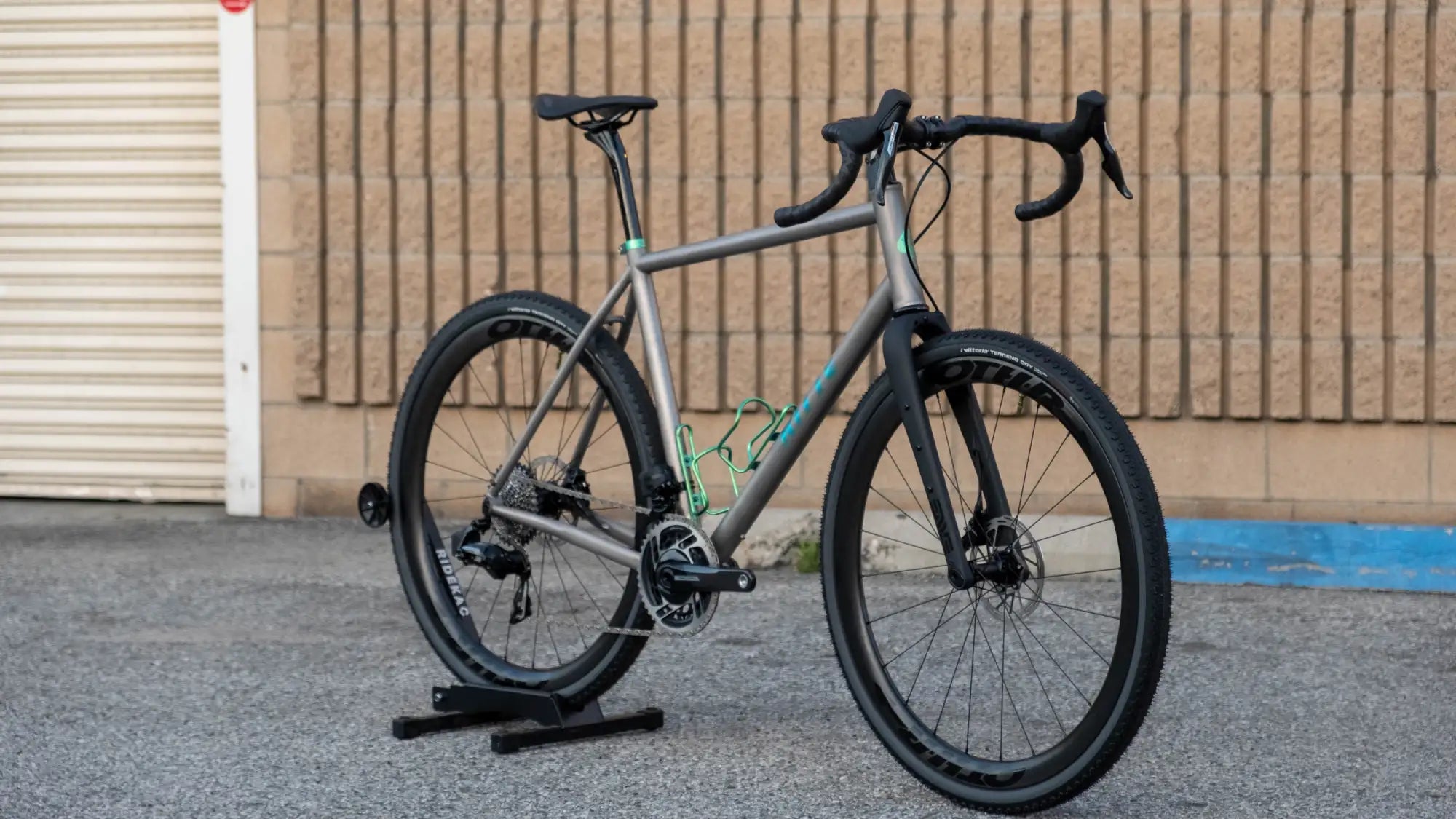 A sleek matte gray gravel bike with black handlebars and tires, featuring a vibrant teal accent on the frame and matching green water bottle cage.