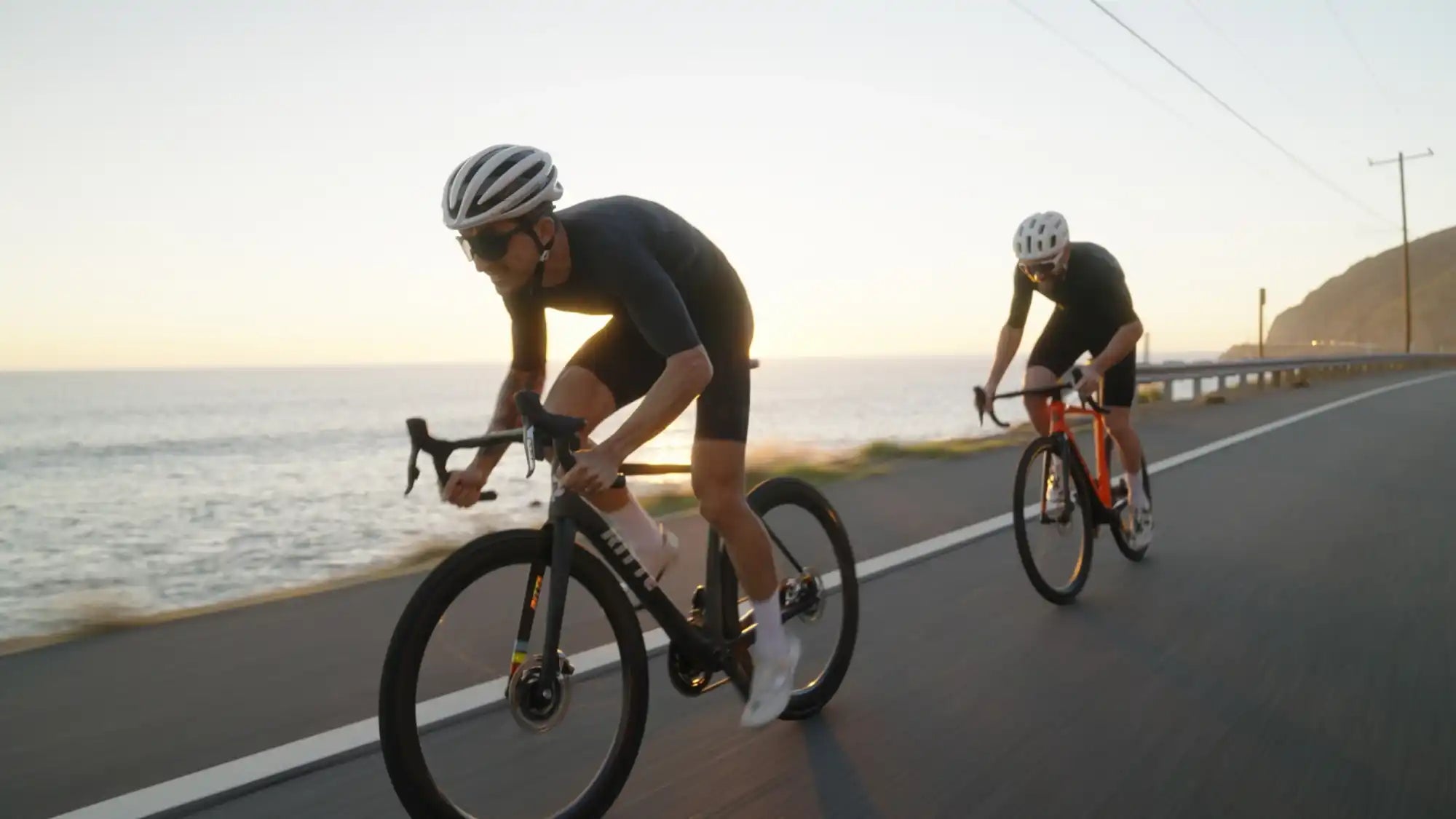 Two cyclists riding road bikes along a coastal highway at sunset.