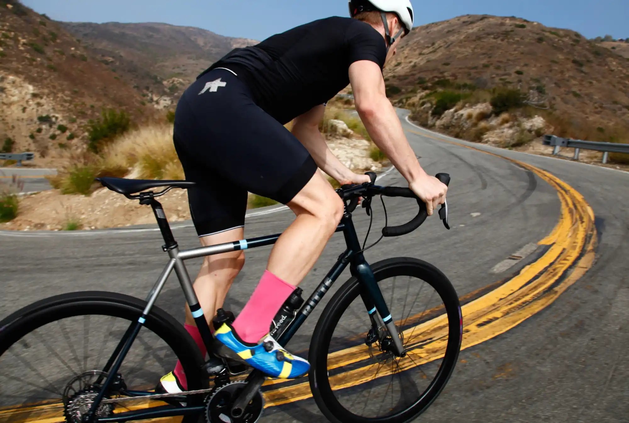 Cyclist wearing black shorts and bright pink socks riding a road bike on a curved road.