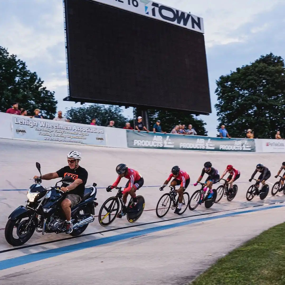 Cyclists racing on a velodrome track with a motorcycle pacer leading them.