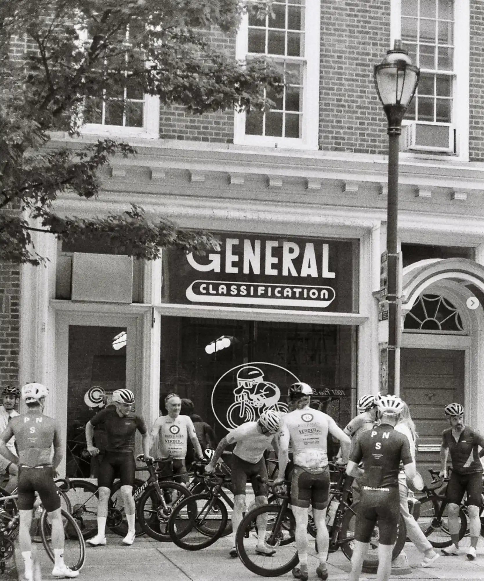 Group of cyclists gathered in front of a ’General Classification’ storefront.